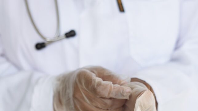 Faceless ethnic medical worker in lab coat and stethoscope taking of transparent gloves after approaching patients for examination in modern hospital